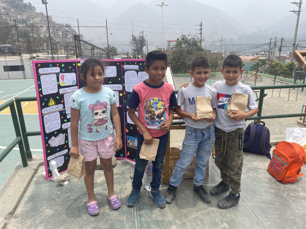 Gruppe von Kindern auf einem Basketballfeld mit Papiertüten in den Händen, Tüten und Gegenstände rechts und eine Tafel mit Text links, vor einer Außenkulisse mit Zaun, Pfosten, Straßenlaternen, Bäumen, Gebäuden, Hügeln und Himmel.