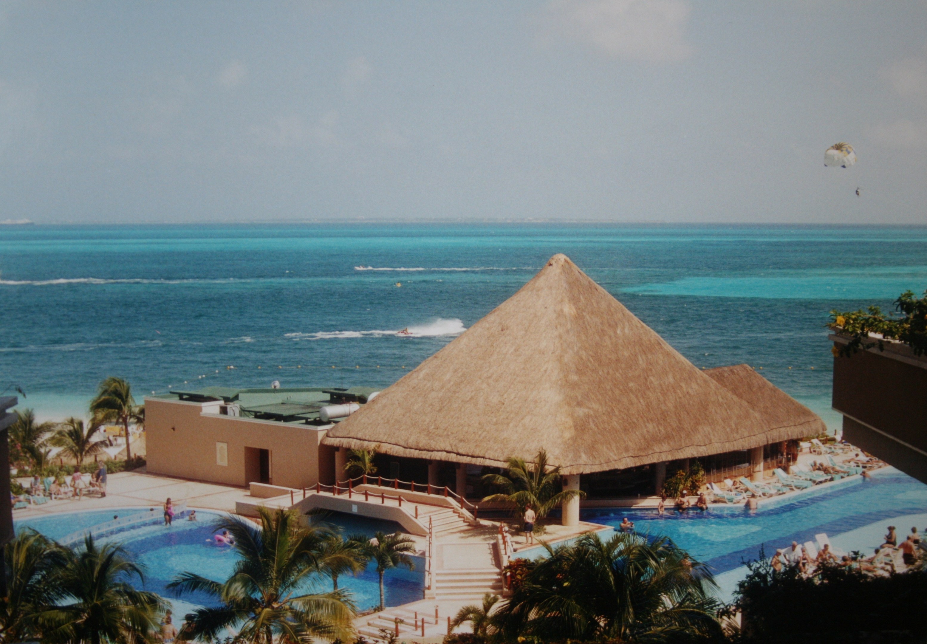 Ein Resort mit einem großen Swimmingpool, umgeben von Palmen, Hütten und Treppen, mit Menschen, die schwimmen, und einem Ozean, einer bewölkten Himmel und einem Fallschirm im Hintergrund.