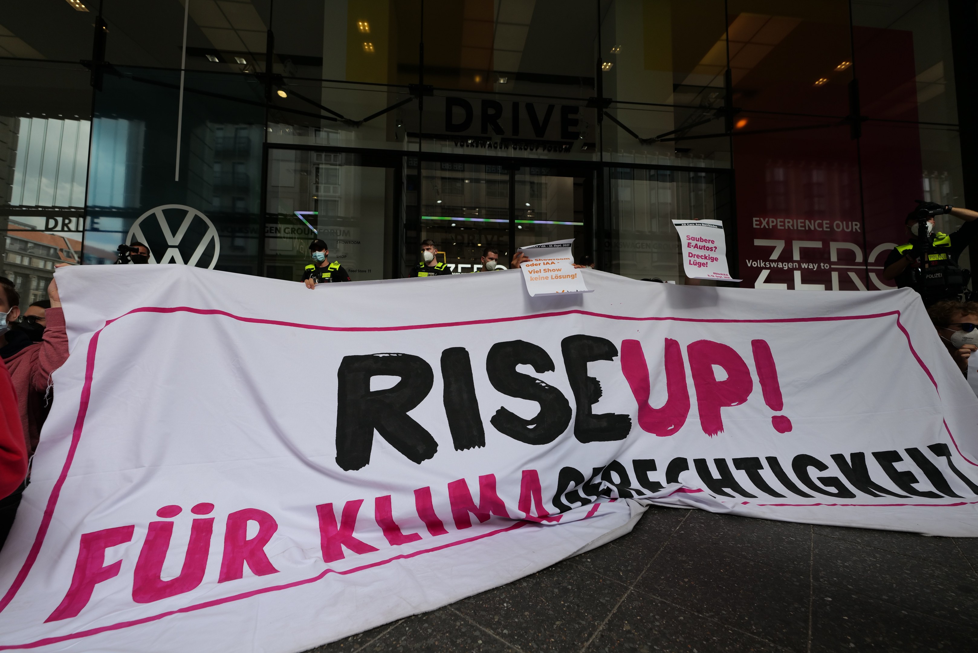 Menschen mit Masken halten ein "Aufstehen"-Schild und Plakate vor einem Glasgebäude bei Nacht.