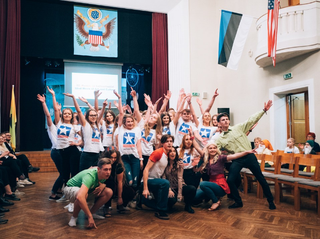 Eine Gruppe von Menschen posiert für ein Foto auf einem nationalen Jugendkongress-Event vor sitzenden und stehenden Zuschauern, mit einem Anzeigebildschirm, Vorhängen, Flaggen, einem Schild, einer Tür, einem Geländer und Deckenlampen im Hintergrund.