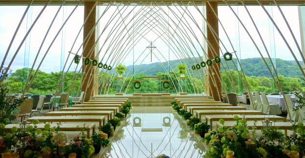 Eine Hochzeitszeremonie in einem großen, geschmückten Saal mit Tischen in halbrunder Anordnung, umgeben von Blumenarrangements, Bäumen, Hügeln und einem klaren blauen Himmel.