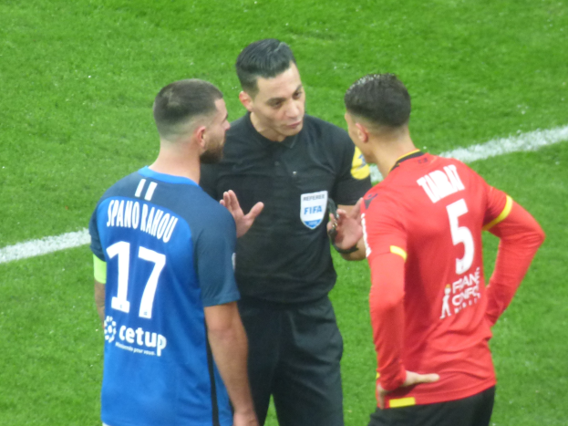 Schiedsrichter spricht mit zwei Männern auf einem Fußballfeld, einer in schwarzer Hose und Hemd, der andere in einem blauen und roten T-Shirt mit Text, beide tragen Brillen.