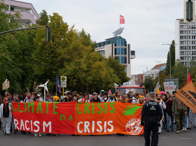 Eine Gruppe von Menschen marschiert auf einer Straße und hält ein "Klima-Krise ist eine Krise"-Schild hoch, mit Bäumen, Laternenmasten und Schildern an der Straße, einem geparkten Fahrzeug in der Nähe, Gebäuden im Hintergrund und einem klaren blauen Himmel.