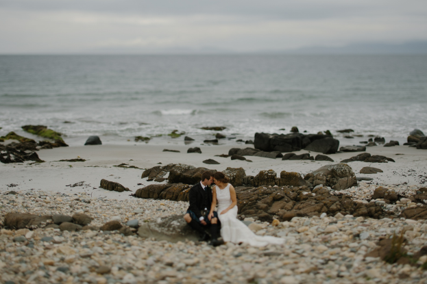 Bräutigam und Braut umarmend und küssend auf einem felsigen Strand mit dem Ozean und dem Himmel im Hintergrund.