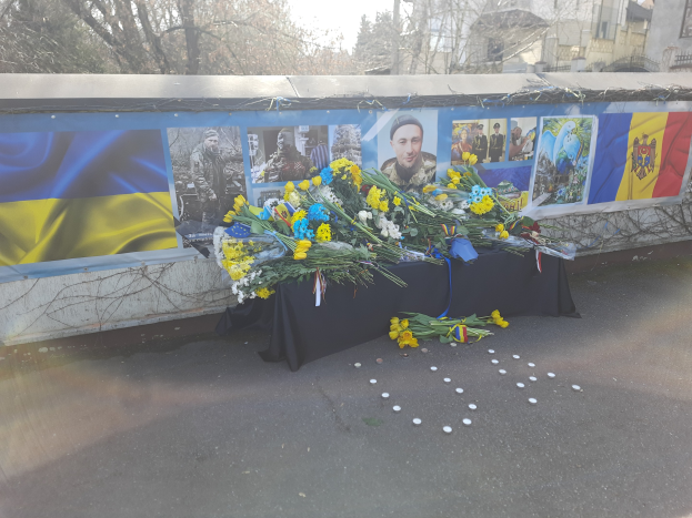 Eine improvisierte Gedenkstätte in Kiew, Ukraine, mit Blumensträußen auf der Straße, Fotos und Flaggen an einer Wand, sowie Bäumen, Gebäuden und einem klaren blauen Himmel im Hintergrund.
