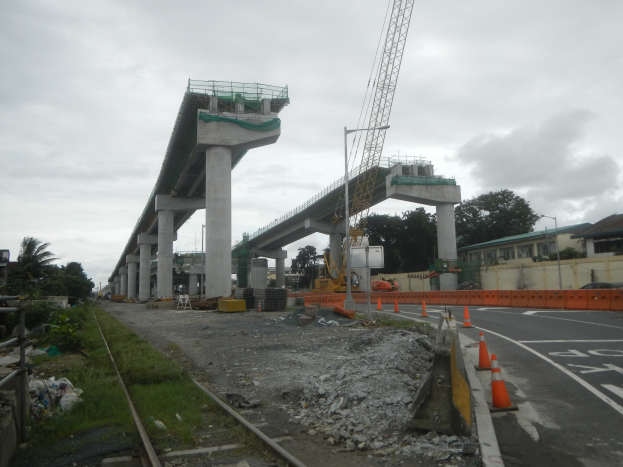 Baustelle mit Brücke im Hintergrund, Straße mit Verkehrskegeln markiert, Bahnschiene links, verstreute Steine und Gras, Bäume und Gebäude auf beiden Seiten und ein bewölkter Himmel.