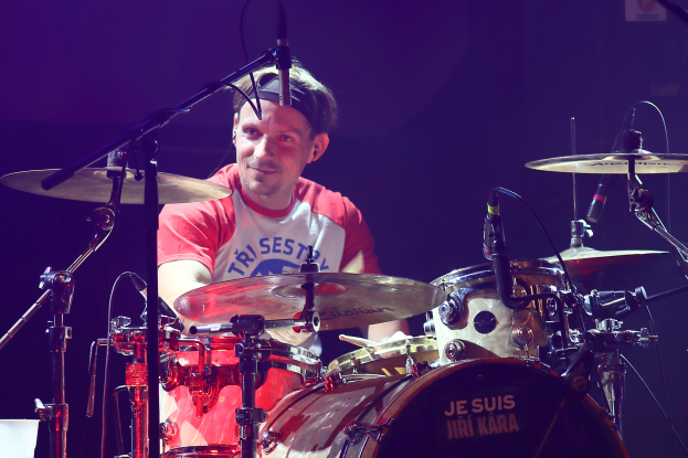 Ein Mann, der auf der Bühne Schlagzeug spielt, trägt ein rotes und weißes T-Shirt und lächelt, umgeben von mehreren Mikrofonen.