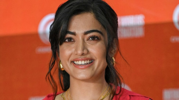 Frau in rotem Kleid mit elegantem Dutt und dezentem Make-up, die in die Kamera lächelt, vor einem gelben Banner mit der Aufschrift 'Die besten Schönheits Looks von Sonam Kapoor beim Cannes Film Festival.'