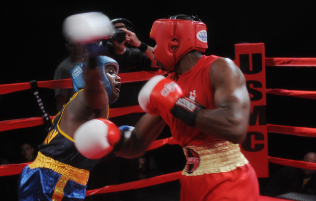 Zwei Boxer kämpfen im Ring, während ein Fotograf und Zuschauer im Hintergrund zu sehen sind, unter dramatischer Beleuchtung.