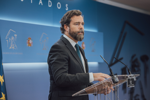 Ein Mann in Anzug und Krawatte steht an einem Podium mit zwei Mikrofonen vor sich, links ein Fahne und hinter ihm eine Wand mit einer Aufschrift.