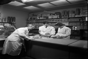 Drei Männer in weißen Kitteln stehen an einem Tresen in einer Apotheke mit Papieren und Gegenständen darauf, mit Regalen im Hintergrund, die mit verschiedenen Gegenständen gefüllt sind, dargestellt in Schwarz-Weiß.