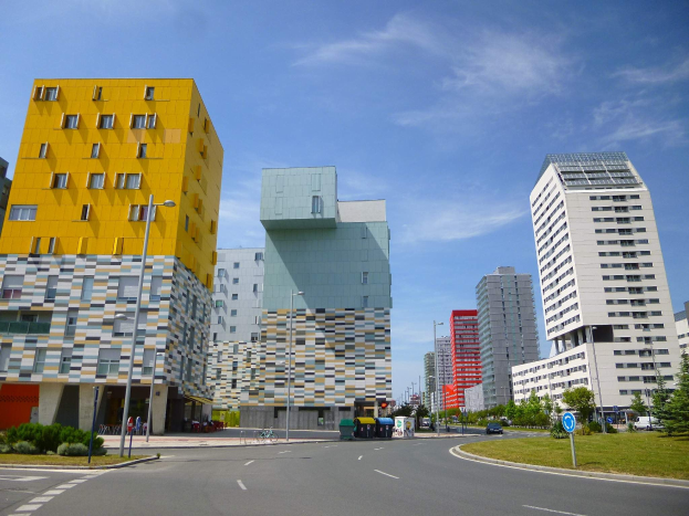 Eine Straße in der Stadt mit hohen Gebäuden im Hintergrund, Straßenmasten, Straßenlaternen, Kraftfahrzeugen, Infotafeln, Büschen, Bäumen und einem bewölkten Himmel, mit Architektur der Universität Rotterdam.