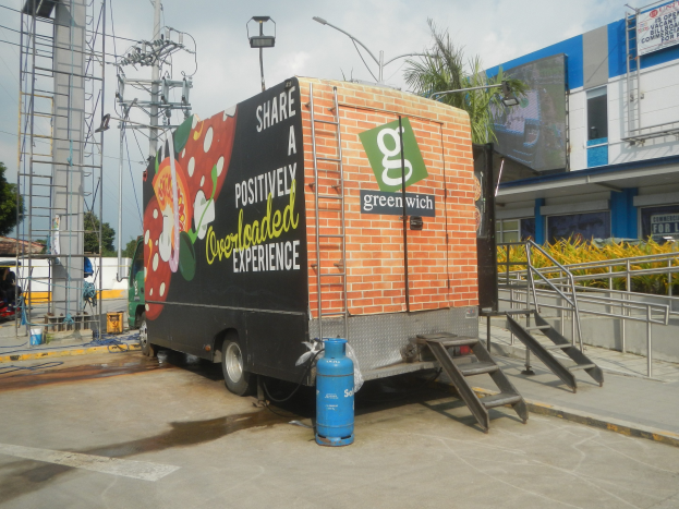 Ein weißer Lkw vor einem Gebäude mit einer Tafel, auf der "Teilen Sie ein positiv überladenes Erlebnis" steht, mit Stufen, einem Zylinder auf dem Boden, Geländern, Strommasten mit Drähten, Laternen, Bäumen und einem bewölkten Himmel im Hintergrund.