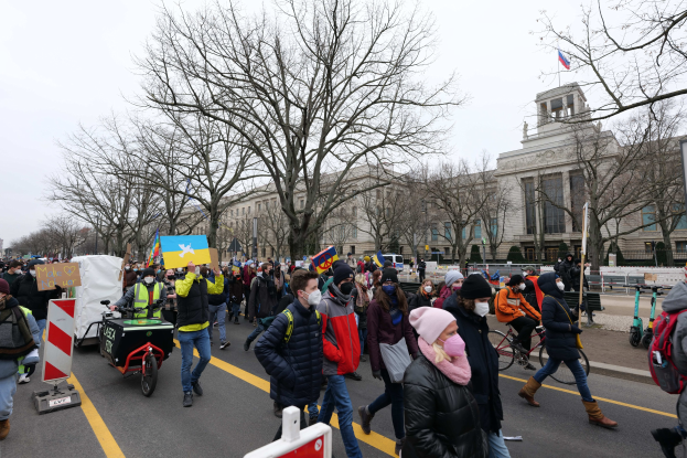 Eine große Gruppe von Menschen bei einer Demonstration auf einer Stadtstraße, einige halten Schilder und andere fahren Fahrräder, mit Bäumen und einem Gebäude im Hintergrund unter einem klaren blauen Himmel.