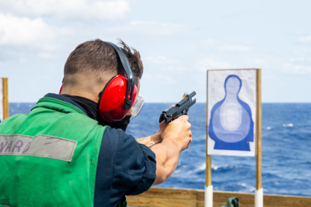 Ein Mann in einer grünen Jacke und roten Ohrschützern schießt mit einer Pistole auf eine Zielscheibe, vor einem hölzernen Geländer mit zwei Brettern stehend, mit einem Meer und einem klaren blauen Himmel im Hintergrund.