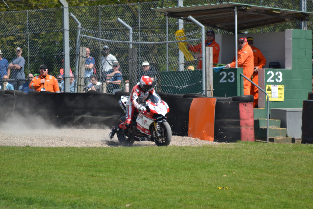 Ein Motorradfahrer in Helm und Handschuhen fährt über ein grasbewachsenes Feld mit Zuschauern, Absperrungen, Treppen und einem nummerierten Schuppen im Hintergrund.