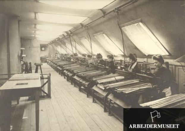 Schwarz-weißes Foto von Frauen, die an Textilmaschinen in einer Fabrik arbeiten, sitzend an Tischen mit einer Uhr an der Wand im Hintergrund.