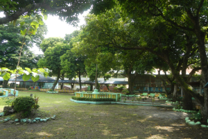 Ein Park mit gr├╝nem Gras, Pflanzen und B├Ąumen, mit einem zentralen Brunnen und einem klaren blauen Himmel im Hintergrund.
