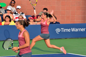 Zwei Frauen halten Tennisrackets und stehen vor einer Wand.