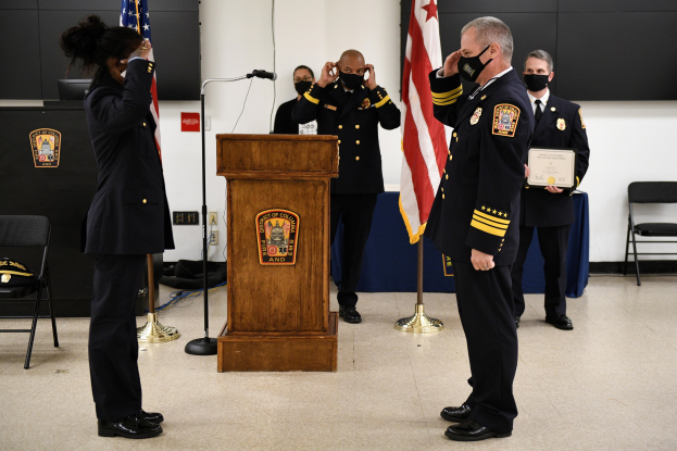 Gruppe uniformierter Personen in Masken vor einem Podium mit Mikrofon, die eine Urkunde während einer Vereidigung eines neuen Feuerwehrchefs halten, umgeben von Flaggen, Stühlen, Tischen und einer Wand.