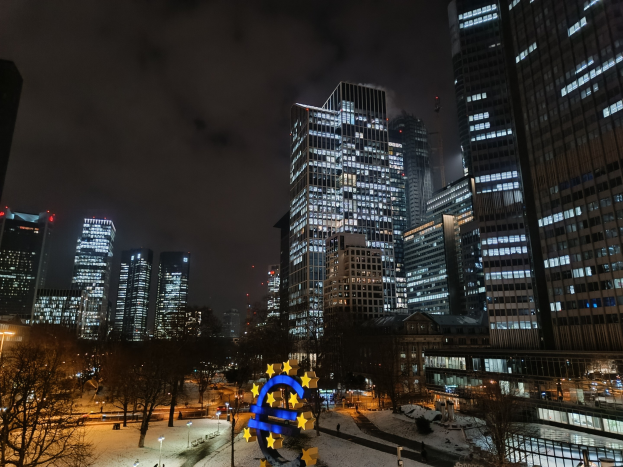 Europäischer Zentralbank-Hauptsitz in Frankfurt, Deutschland bei Nacht, mit beleuchteten Gebäuden, Bäumen, Straßenlaternen, Lichtern und Fußgängern unter einem bewölkten Himmel.