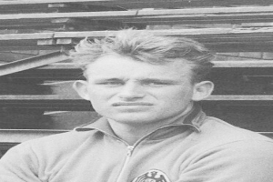 Ein Schwarz-Weiß-Foto eines älteren Mannes in einem Jackett vor einem Stapel Holzplanken.