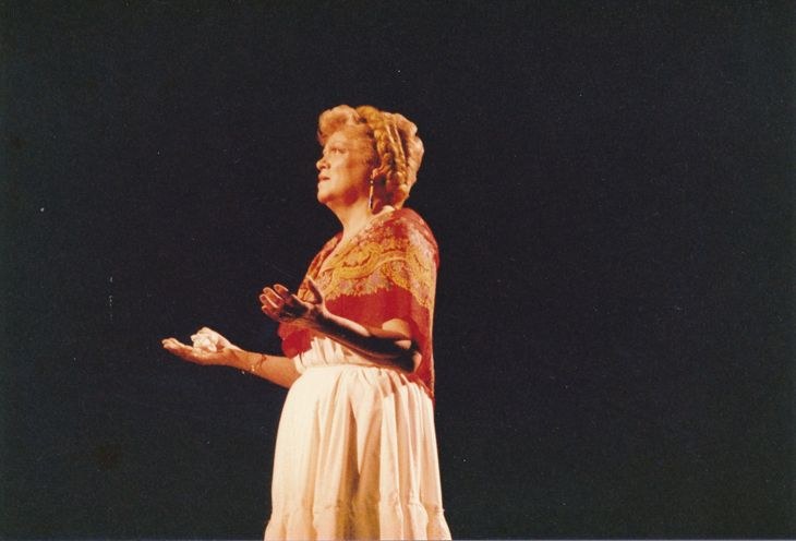 Eine Frau in einem weißen, hochgeschlossenen, langärmeligen Kleid steht auf einer dunklen Bühne mit ausgestreckten Armen, ihr Haar ist zu einem Dutt gebunden und ihr Gesichtsausdruck ernst.