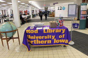 Eine Ausstellung mit einem Tisch, auf dem verschiedene Landesflaggen, eine Box, Papiere und ein Banner liegen, eine Kugel links daneben, eine Mitteilungstafel rechts daneben und mehrere Tische und Stühle in der Ferne.