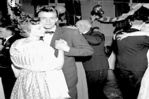 Ein Schwarz-Weiß-Foto von einem Mann und einer Frau, die auf einer Party tanzen, umgeben von lachenden Zuschauern und festlichen Dekorationen und Ballons im Hintergrund.