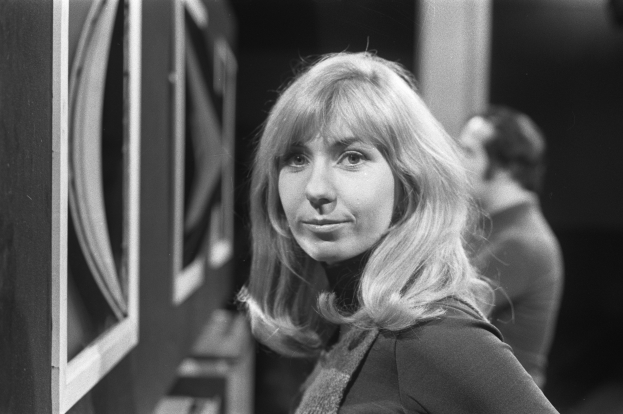 Frau in Kleid vor Fernseher mit einer weiteren Person dahinter, in einem Schwarz-Weiß-Setting mit leicht unscharfem Hintergrund.