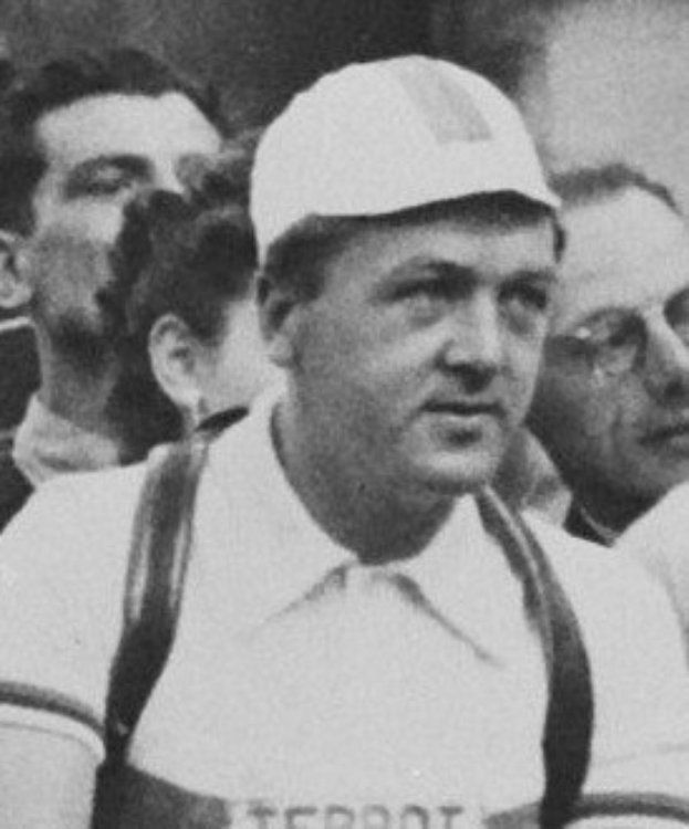 Schwarzes und weißes Foto eines Mannes mit Mütze und Hosenträgern, der vor einer Gruppe von Menschen steht, die mutmaßlich ein legendärer Fußballspieler ist.