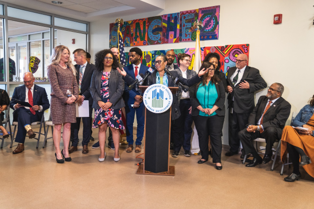 Gruppe von Menschen vor einem Podium mit Mikrofon und einer Tafel mit der Aufschrift "Sancho Stadtrat tritt dem Stadtrat bei", mit sitzenden und stehenden Zuschauern, Flaggen, Feuerlöscher und Deckenleuchten im Hintergrund.