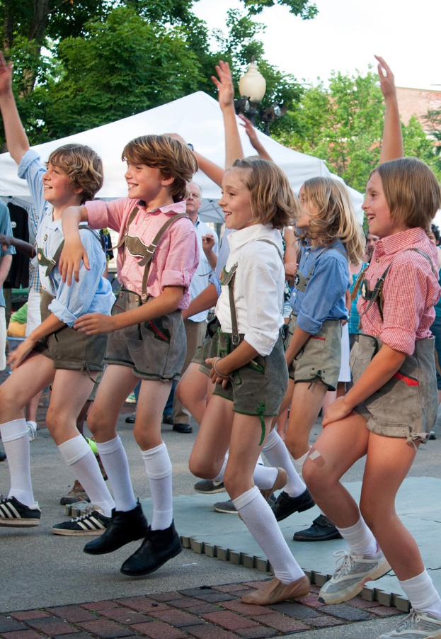 Kinder in traditioneller bayrischer Tracht tanzen auf einer Straße während eines Outdoor-Events, umgeben von einer Menge, Zelten und Gebäuden unter einem klaren blauen Himmel.