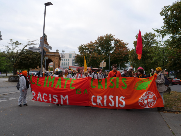 Gruppe von Menschen, die eine Straße entlanggehen und ein 'Klimakrise ist eine Krise'-Plakat halten, mit Fahrzeugen, Gebäuden und einem klaren blauen Himmel im Hintergrund.