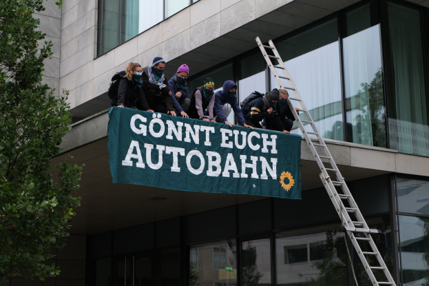 Eine Gruppe von Menschen steht auf einem Gebäude und hält eine Fahne, mit einem Baum auf der linken Seite und Glasfenstern im Hintergrund, die scheinbar für die deutsche Regierung protestieren.