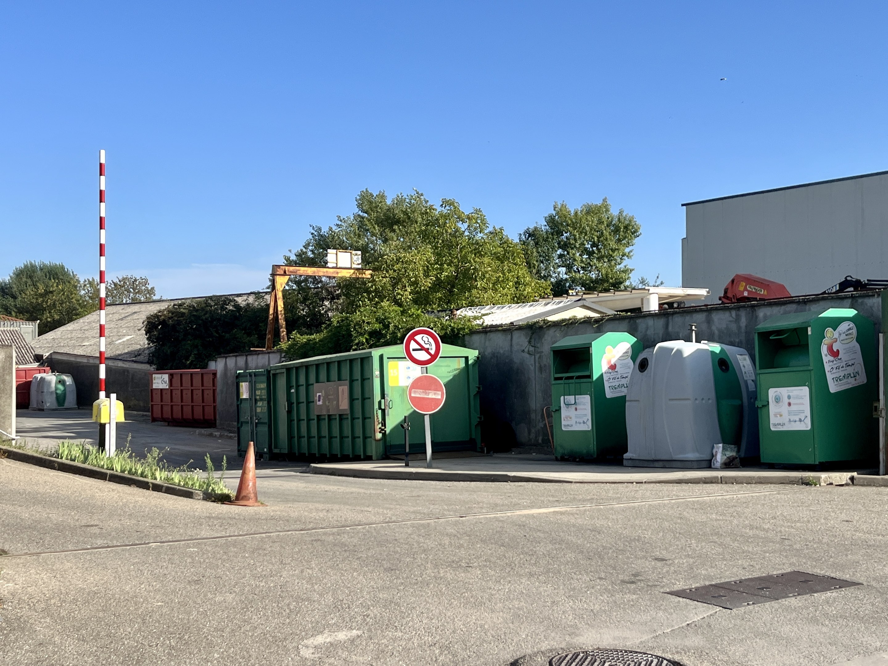 Grüne und weiße Mülltonnen entlang einer Parkplatzseite aufgereiht, mit einem Verkehrskegel im Vordergrund und einer Hinweistafel auf einem Pfahl im Hintergrund unter einem bewölkten Himmel mit Bäumen und Gebäuden in der Ferne.