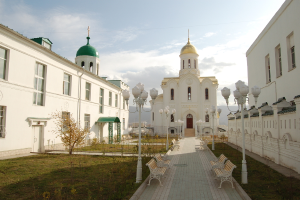 Eine Stadtansicht mit Gebäuden mit sichtbaren Fenstern, einer grasbewachsenen Fläche mit einem Lichtmast, Blumenkübel und Bänken, einem Fußweg und einem bewölkten Himmel.