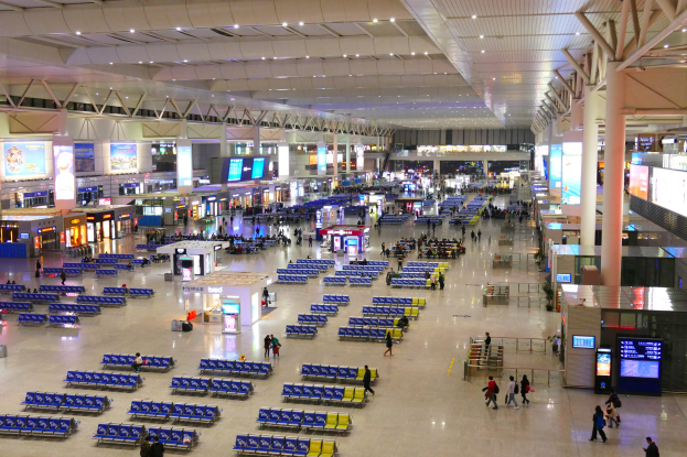Ein belebter Flughafen-Terminal-Innenraum mit Menschen, blauen Stühlen, Geschäften, Texttafeln, Bildschirmen, Säulen, Lampen und Stangen.