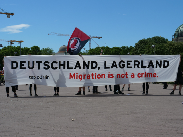 Eine Gruppe von Menschen mit einem Transparent, auf dem "Deutschland, Lagerland Migration ist kein Verbrechen" steht, und einer Flagge, im Hintergrund Straßenmäste, Lichter, Bäume, Kräne, ein Gebäude und ein bewölkter Himmel.