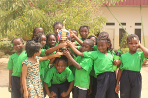 Gruppe von Kindern in einer Schulumgebung mit einem Pokal, umgeben von Bäumen und einem Gebäude