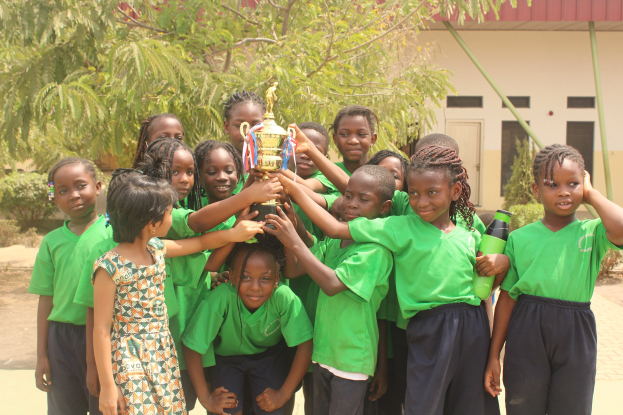Gruppe von Kindern in einer Schulumgebung mit einem Pokal, umgeben von Bäumen und einem Gebäude