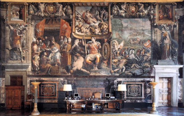 Großer Raum mit einem prominenten Gemälde an der Wand, ein Tisch mit Gegenständen, zwei Lampen, zwei Türen und ein Teppich, befindet sich im Palazzo della Signoria in Florenz, Italien.