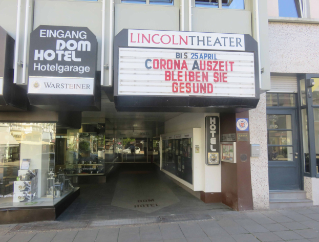Außenansicht des Lincoln Theaters in Berlin, Deutschland, mit Glasfenstern und -türen sowie einer Tafel mit Text und einem Innenraum, der eine pulsierende Stadtlandschaft suggeriert.