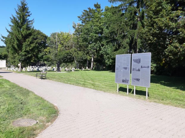 Ein Park mit einem zentralen Gras- und Baumgesäumten Weg, der von einer Bank im Vordergrund und Informationsschildern, Gräbern im Hintergrund und einem klaren blauen Himmel umgeben ist.