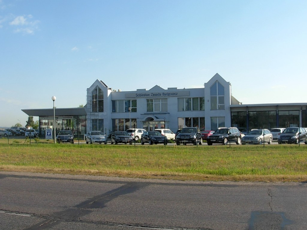 Autohandel mit parkenden Autos, umgeben von Gebäuden, Straßeninfrastruktur, fahrenden Fahrzeugen und Bäumen unter einem bewölkten Himmel.