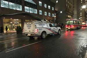 Ein Polizeifahrzeug steht nachts an der Seite einer Stadtstraße, mit Fahrzeugen auf der Straße, Passanten auf dem Gehweg, Topfpflanzen, Straßenlaternen, Gebäuden mit Fenstern und einem bewölkten Himmel.