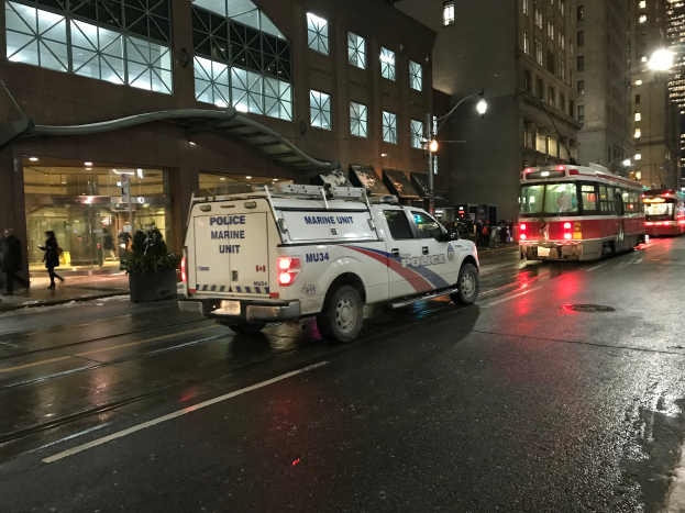 Ein Polizeifahrzeug steht nachts an der Seite einer Stadtstraße, mit Fahrzeugen auf der Straße, Passanten auf dem Gehweg, Topfpflanzen, Straßenlaternen, Gebäuden mit Fenstern und einem bewölkten Himmel.