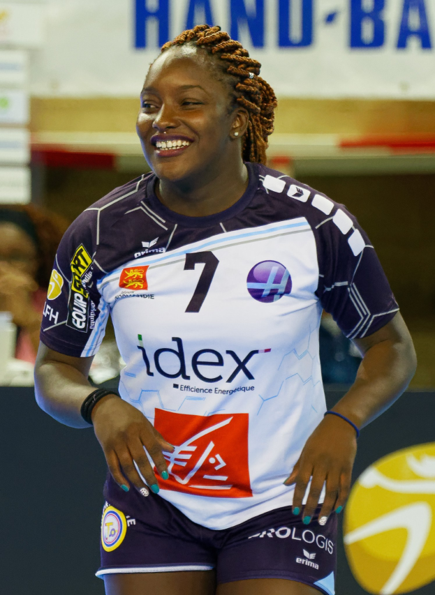 Professionelle Handballspielerin in lila-weißer Uniform, die selbstbewusst auf einem Feld lächelt, mit einem FIVB-Handball-Weltmeisterschaft-2019-Schild im Hintergrund.