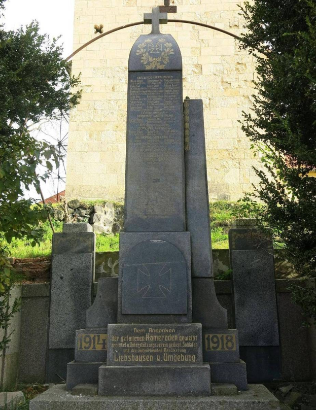 Ein Denkmal aus Stein für deutsche Soldaten, die im Zweiten Weltkrieg starben, steht vor einer Kirche mit einem Kreuz, umgeben von Bäumen und einer Mauer.