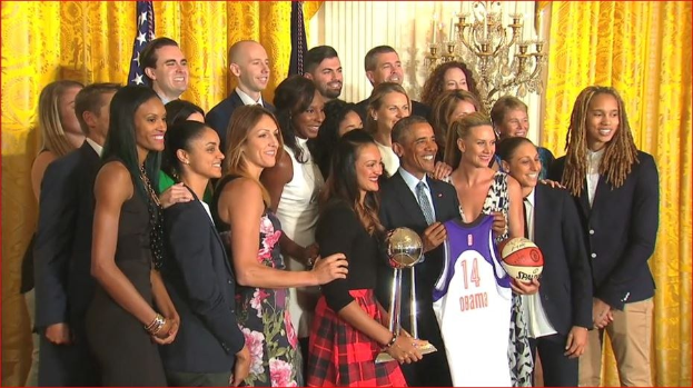 Präsident Obama und First Lady Michelle Obama posieren mit dem Damen-Basketball-Team im Oval Office des Weißen Hauses, halten einen Basketball, Pokal und lächeln mit einer Flagge, Vorhängen und einem Kerzenleuchter im Hintergrund.